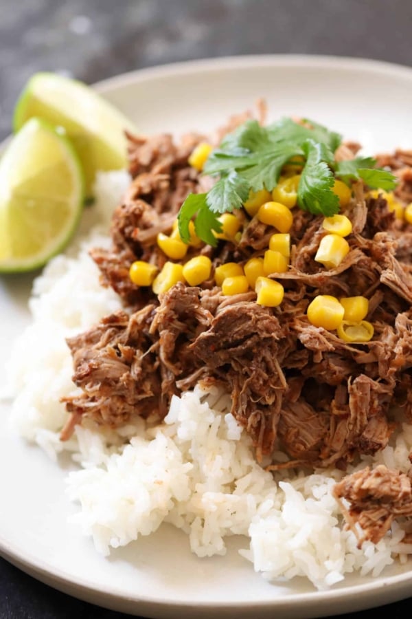 A plate of shredded beef and corn served over white rice, garnished with cilantro and accompanied by lime wedges.