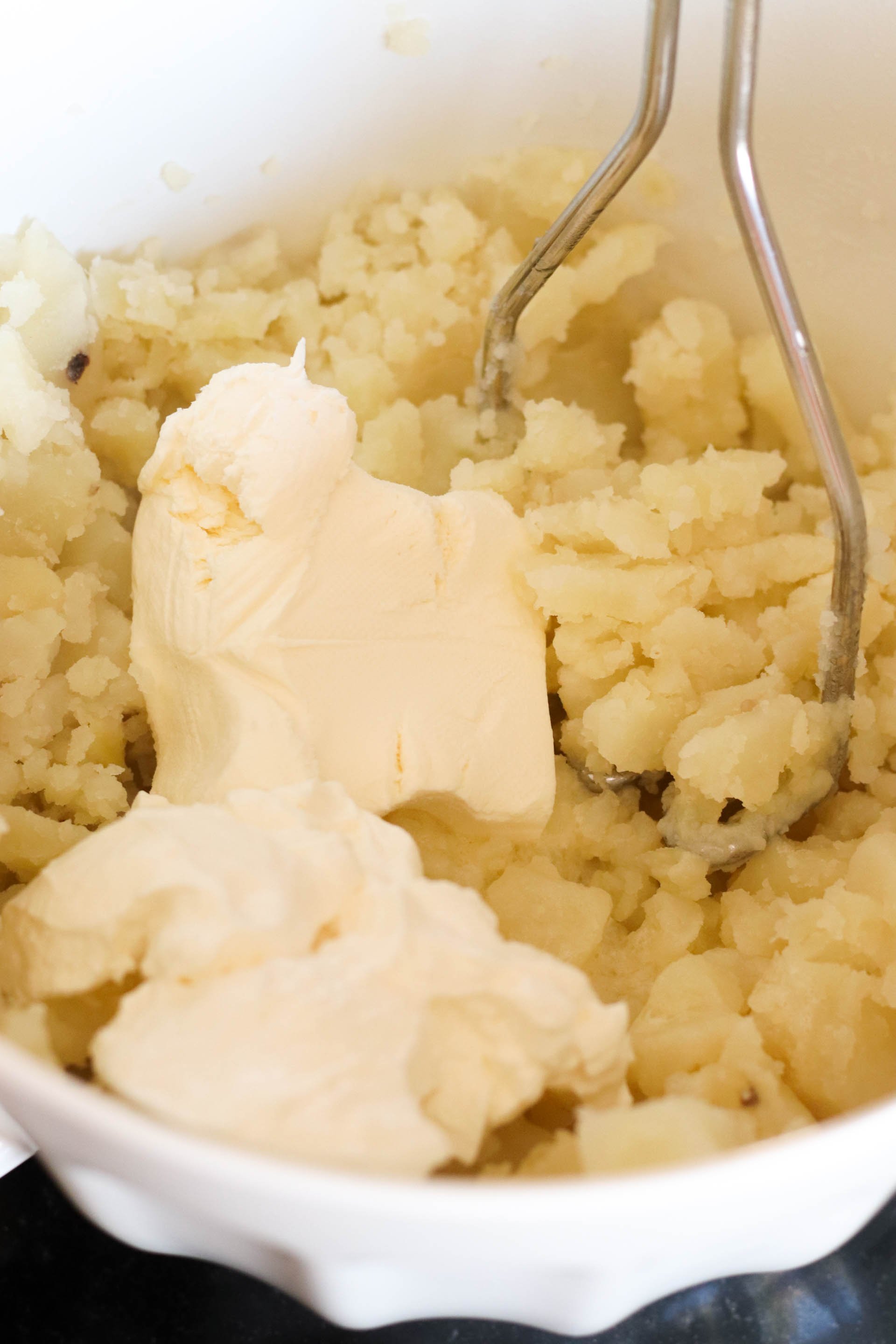 Potatoes, sour cream, and cream cheese being mixed together with a potato masher