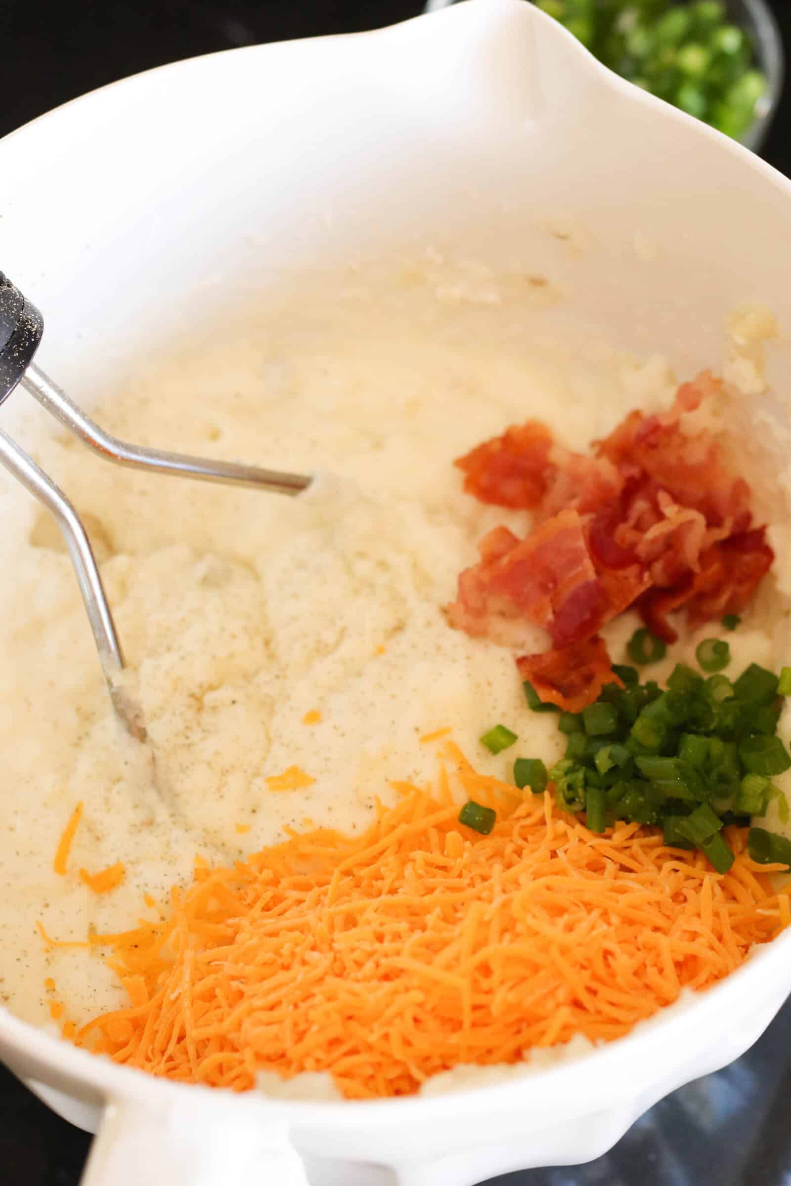 Mashed potato mixture with bacon bits, green onion, and cheese waiting to be mixed in the bowl.