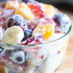 A glass bowl filled with mixed fruit pieces, including bananas, grapes, strawberries, and peaches, coated in a creamy dressing.
