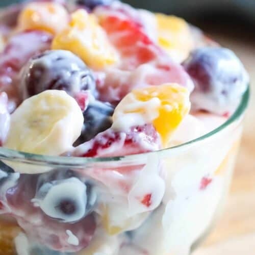A glass bowl filled with mixed fruit pieces, including bananas, grapes, strawberries, and peaches, coated in a creamy dressing.