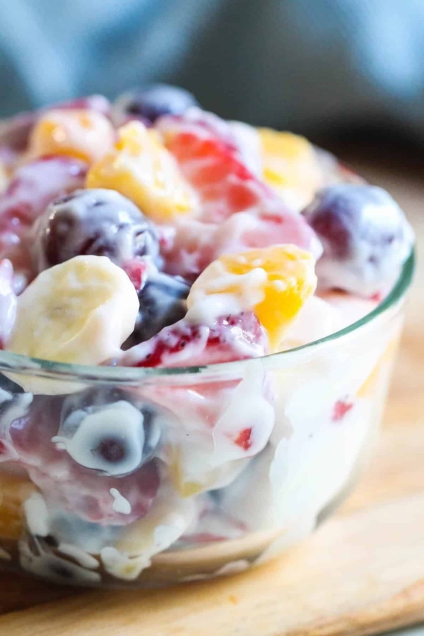 A glass bowl filled with mixed fruit pieces, including bananas, grapes, strawberries, and peaches, coated in a creamy dressing.