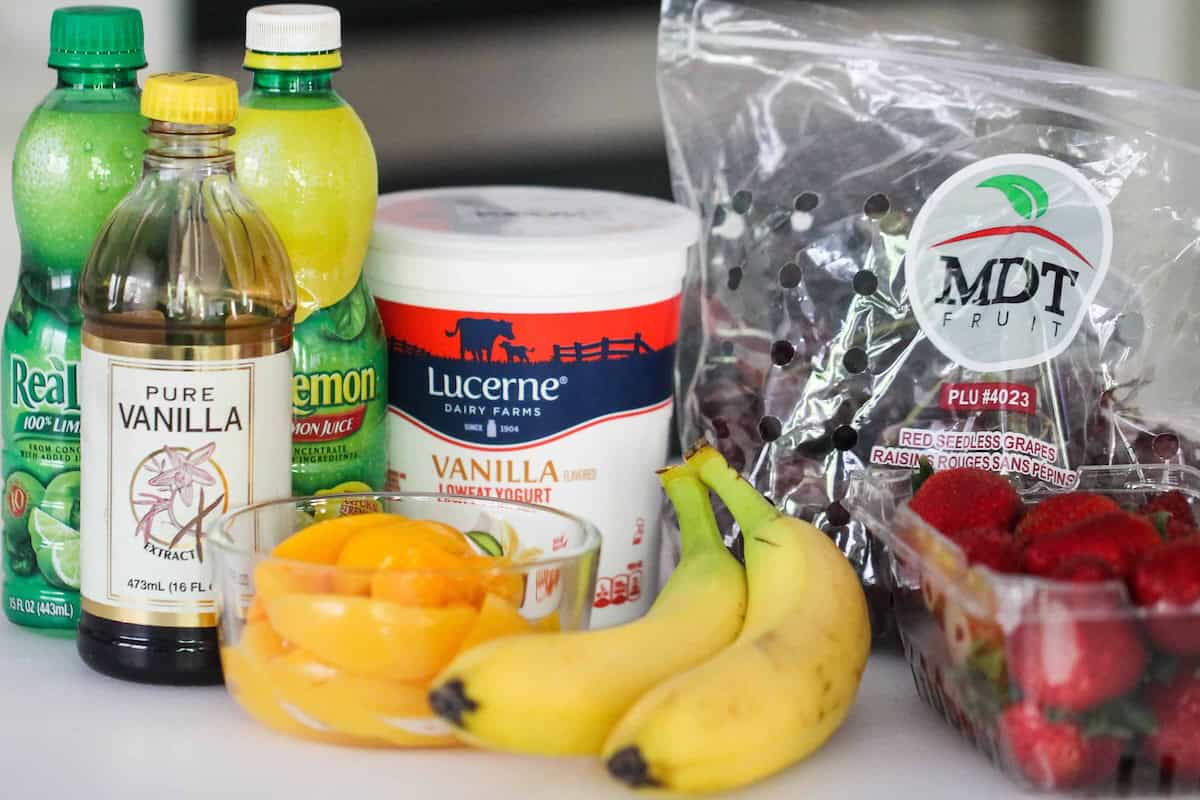 Bottles of lemon juice, vanilla extract, a container of vanilla yogurt, a bowl of canned peaches, bananas, red grapes, and strawberries arranged on a counter.