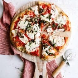 A sliced pizza with melted cheese, mushrooms, and herbs on a wooden board, next to a red-striped cloth and a pizza cutter.