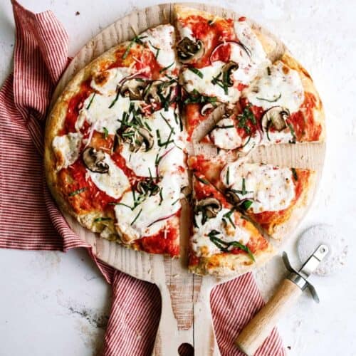 A sliced pizza with melted cheese, mushrooms, and herbs on a wooden board, next to a red-striped cloth and a pizza cutter.