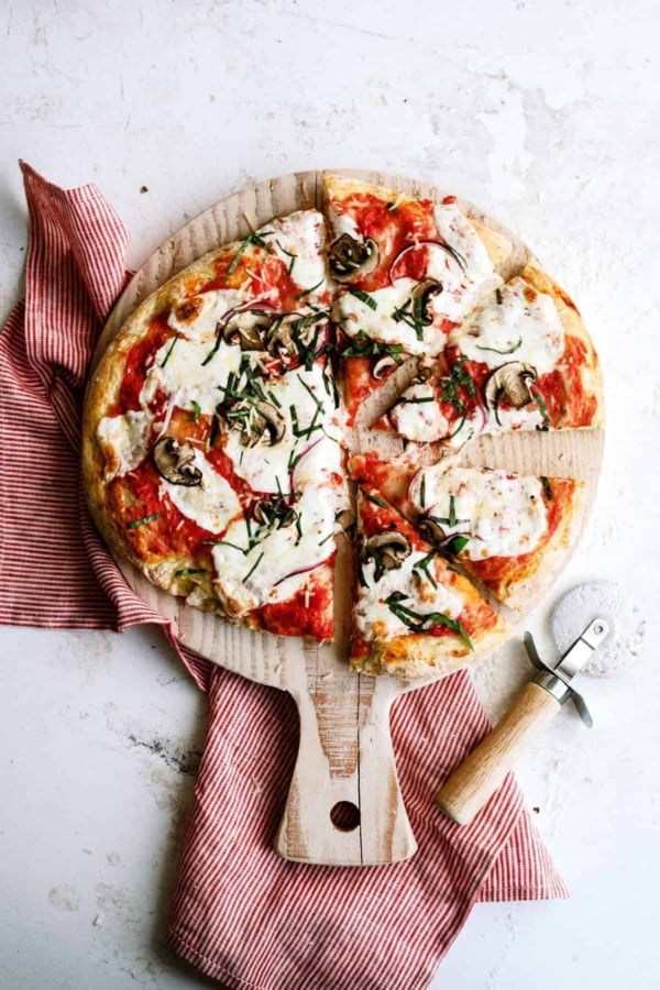 A sliced pizza with melted cheese, mushrooms, and herbs on a wooden board, next to a red-striped cloth and a pizza cutter.