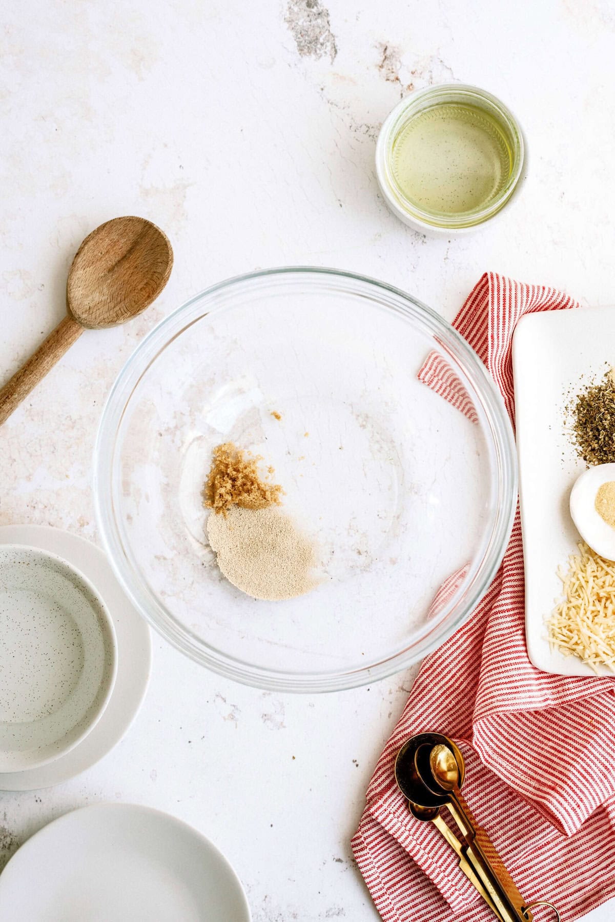 A glass mixing bowl with yeast and sugar inside, surrounded by a wooden spoon, measuring spoons, a small bowl of oil, and various ingredients on a red-striped cloth.