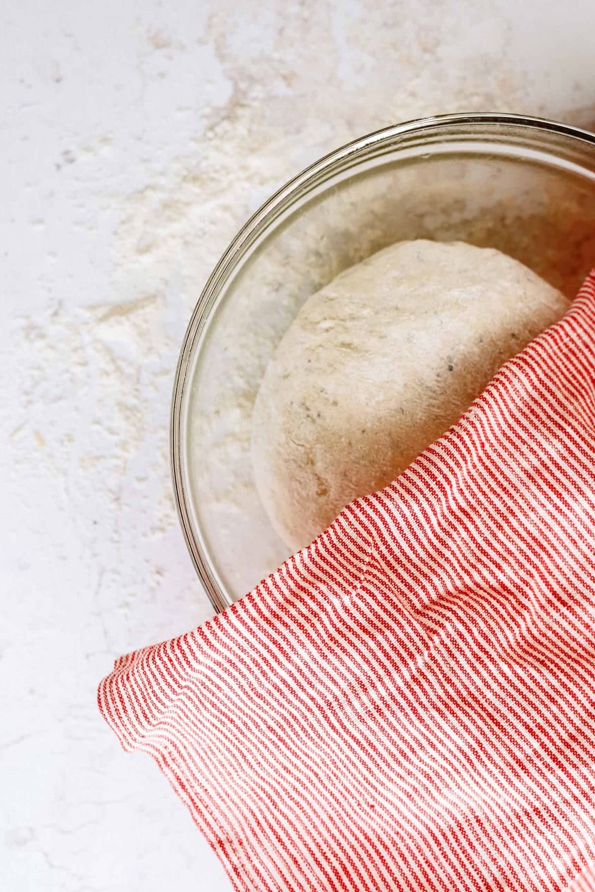 A glass bowl with bread dough partially covered by a red and white striped cloth on a light surface.