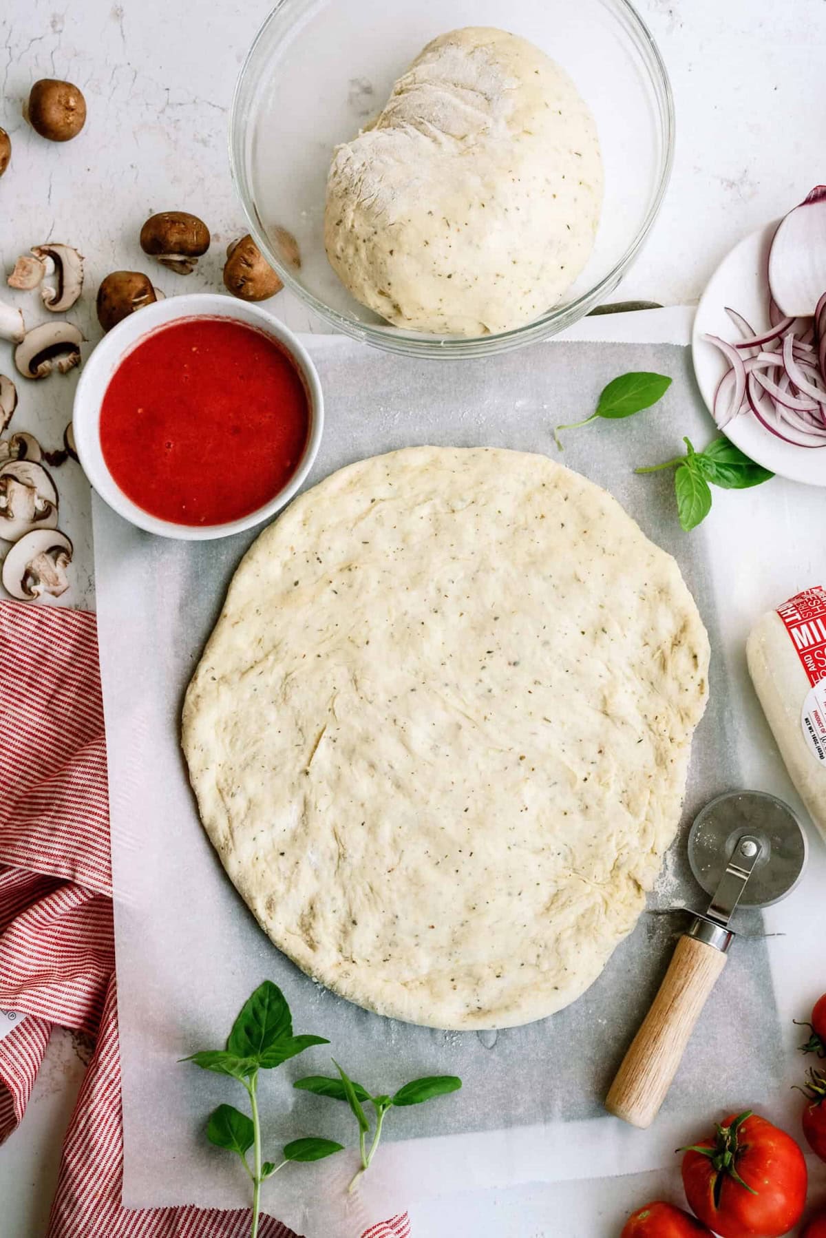 Round pizza dough rolled out on parchment paper, surrounded by pizza sauce, fresh dough, mushrooms, tomatoes, red onion, mozzarella cheese, basil, and a pizza cutter.