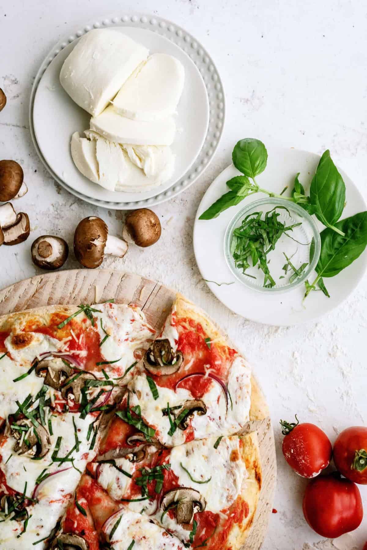 Sliced mushroom and basil pizza on a wooden board, with fresh mozzarella, basil leaves, mushrooms, and tomatoes arranged nearby on a white surface.