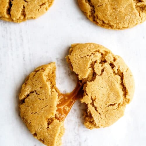 Three cookies on a white surface, with one broken in half revealing a gooey caramel center.