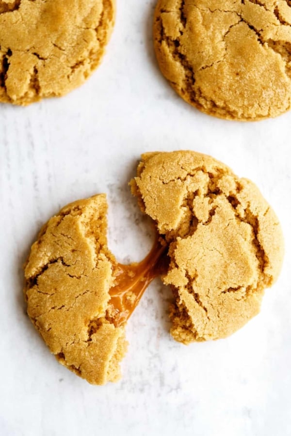 Three cookies on a white surface, with one broken in half revealing a gooey caramel center.