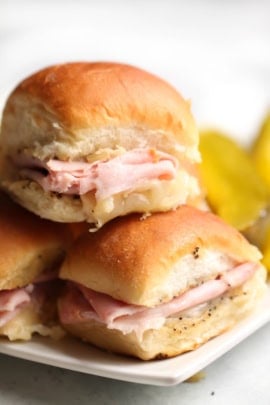 Three sliders with ham and cheese on a white plate, accompanied by pickle slices.