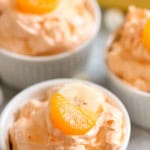 Three small white ramekins filled with an orange-colored creamy dessert, each topped with a slice of mandarin and a banana piece, with a whole banana blurred in the background.