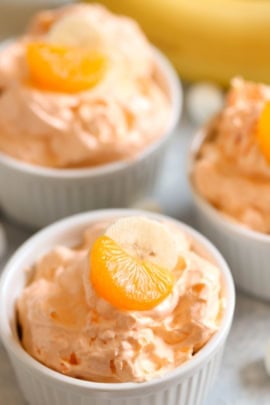 Three small white ramekins filled with an orange-colored creamy dessert, each topped with a slice of mandarin and a banana piece, with a whole banana blurred in the background.