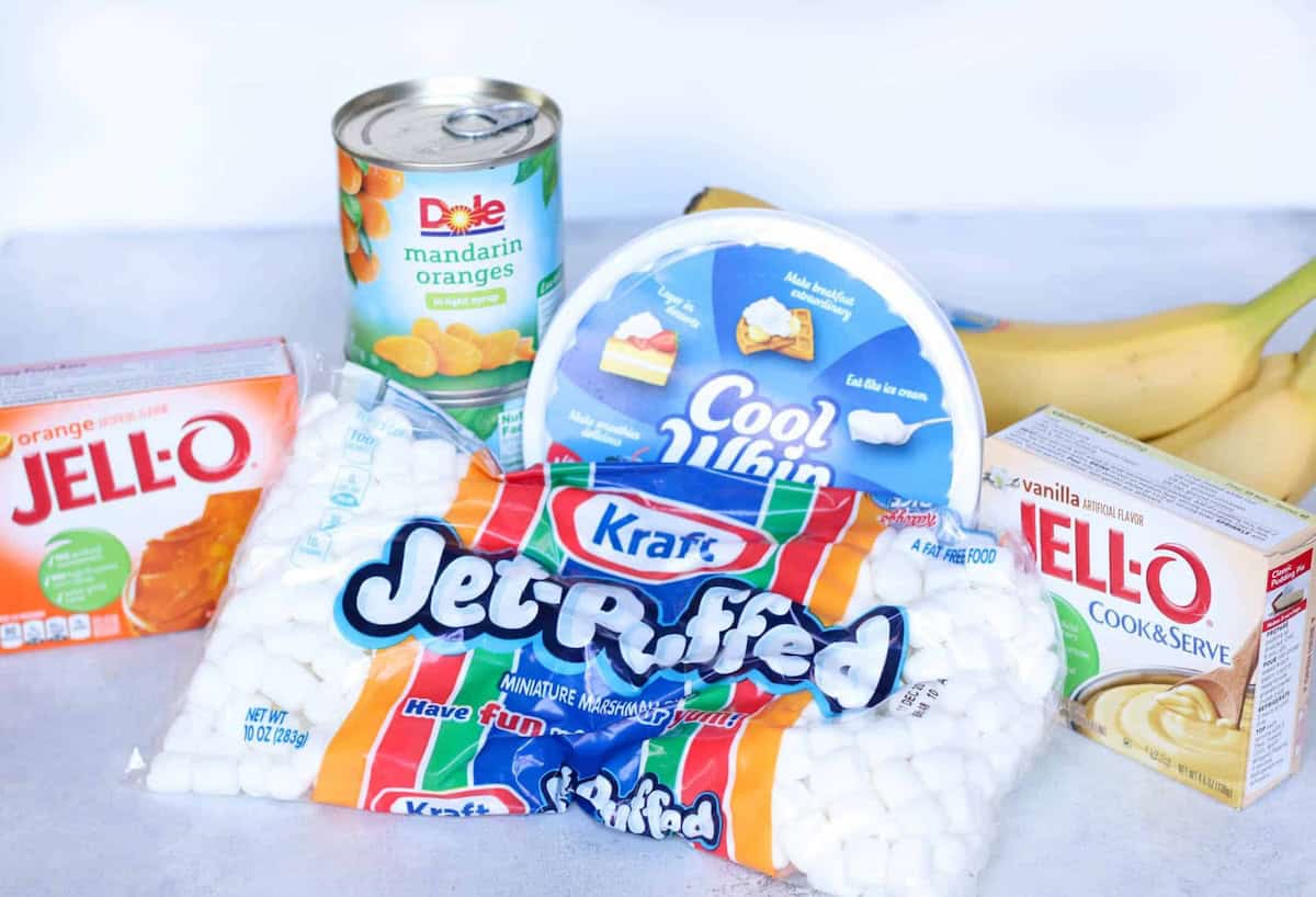 Ingredients on a counter: a can of mandarin oranges, Cool Whip, bananas, orange Jell-O, vanilla pudding mix, and a bag of mini marshmallows.