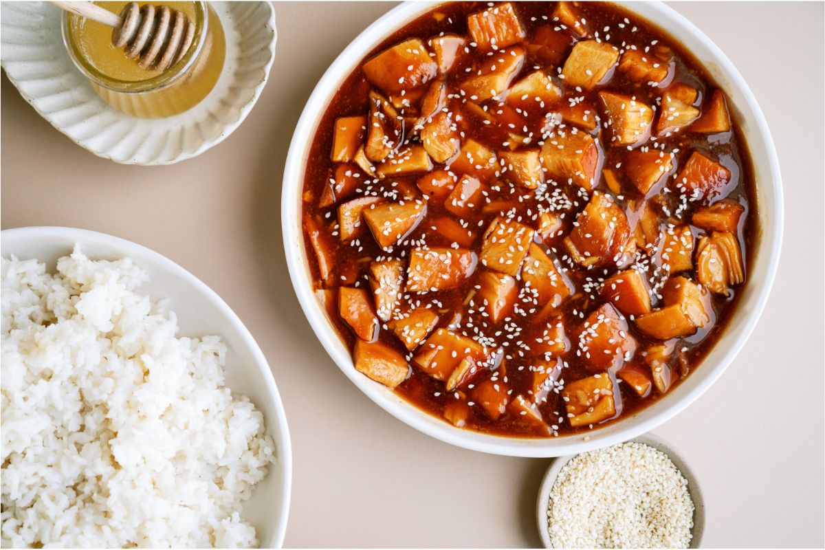 A serving bowl of Slow Cooker Honey Sesame Chicken topped with sesame seeds, a bowl of rice and two small bowls of honey and sesame seeds.