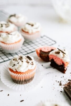 Neapolitan Cupcakes topped with frosting. One on the counter with the cupcake paper removed and one cut in half to show the inside. Other Neapolitan Cupcakes on a cooling rack in the background.