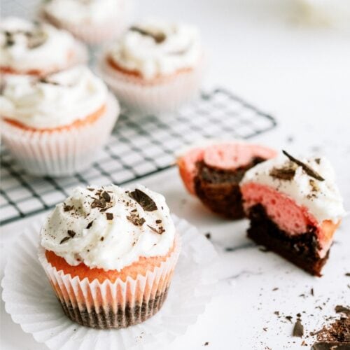 Neapolitan Cupcakes topped with frosting. One on the counter with the cupcake paper removed and one cut in half to show the inside. Other Neapolitan Cupcakes on a cooling rack in the background.