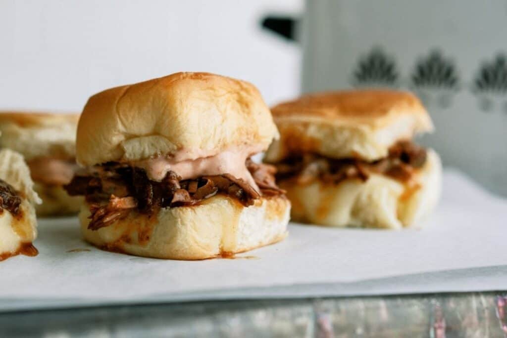 three envelope pot roast sliders with sauce on small buns placed on a white surface, with another slider partially visible in the background.