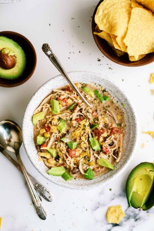 A bowl of chicken tortilla soup topped with diced avocado and surrounded by two spoons and a bowl of tortilla chips. An avocado half and scattered herbs are visible in the background.