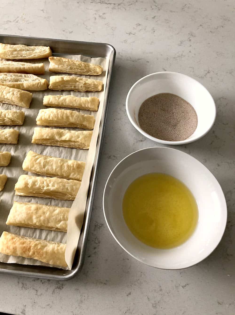 Puff pastry on baking sheet with a bowl of butter and a bowl of cinnamon sugar mix