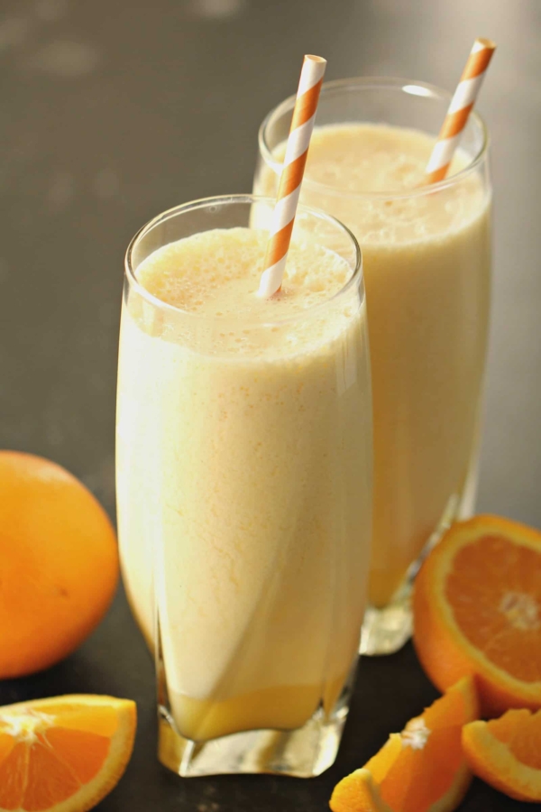 Two glasses of orange smoothies with striped straws, placed next to a whole orange and a sliced orange on a dark surface.