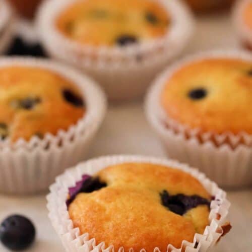 A close-up of freshly baked blueberry muffins in white paper liners, with a few scattered blueberries on a light-colored surface.