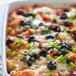 A baked casserole topped with melted cheese, sliced black olives, chopped green onions, and diced red peppers in a white dish.