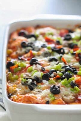 A baked casserole topped with melted cheese, sliced black olives, chopped green onions, and diced red peppers in a white dish.