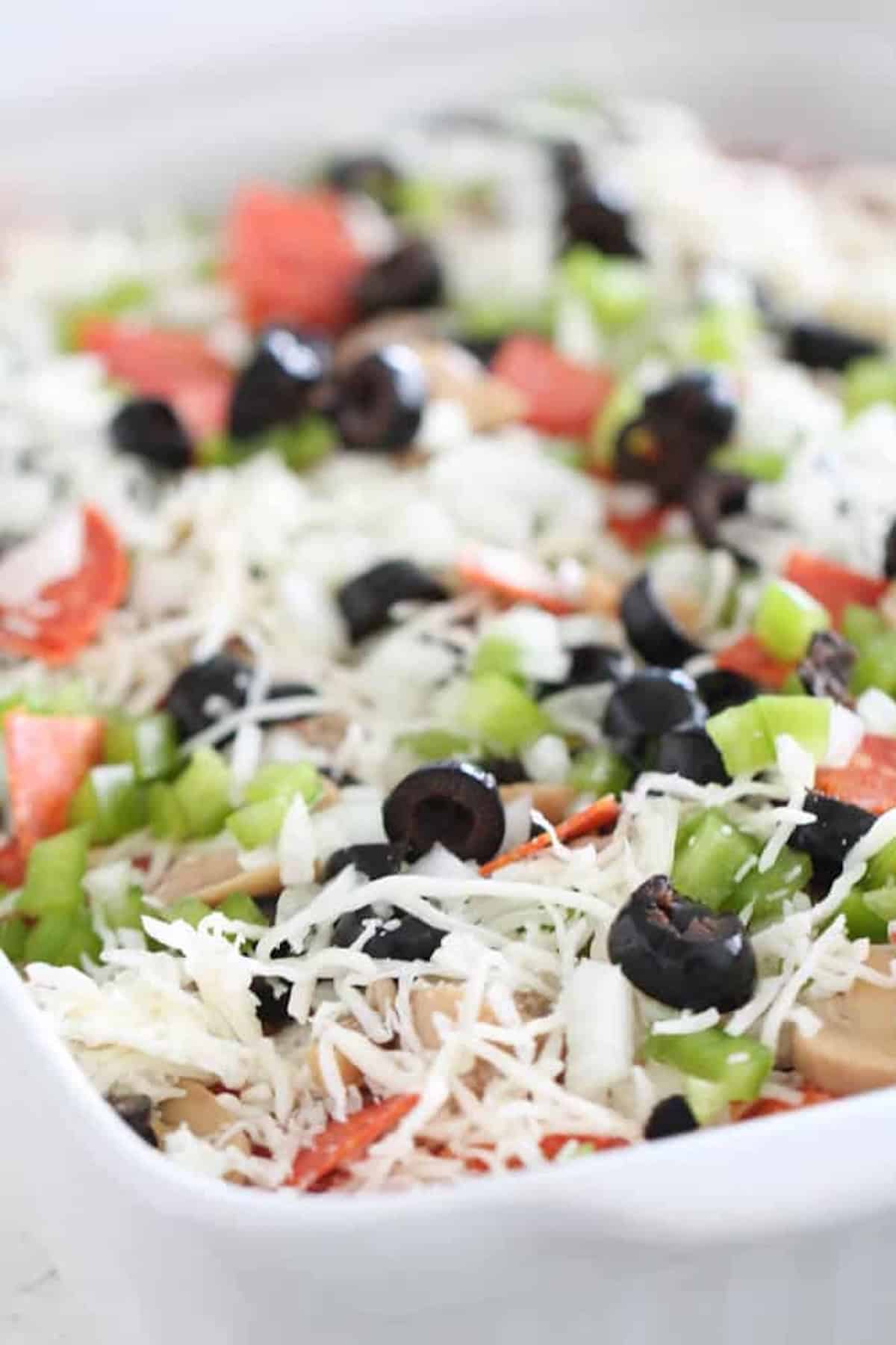 Rectangular dish filled with unbaked pizza casserole topped with shredded cheese, sliced black olives, green bell peppers, onions, mushrooms, and pepperoni.