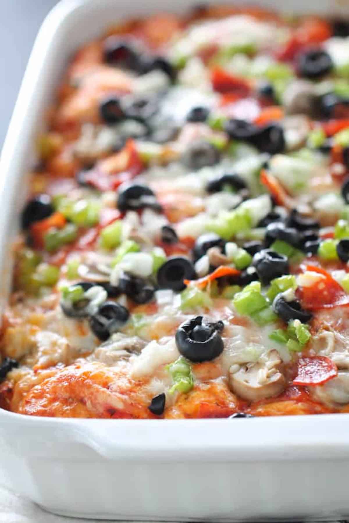 A close-up of a baked casserole topped with melted cheese, black olives, green peppers, mushrooms, and pepperoni slices in a white dish.