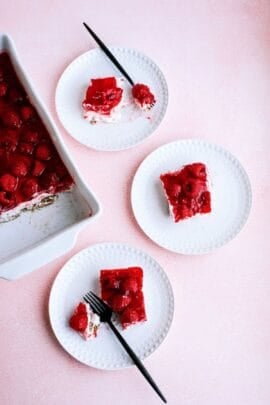 Three white plates with slices of raspberry dessert topped with red jelly, next to a baking dish containing the remaining dessert. Two plates have black forks.