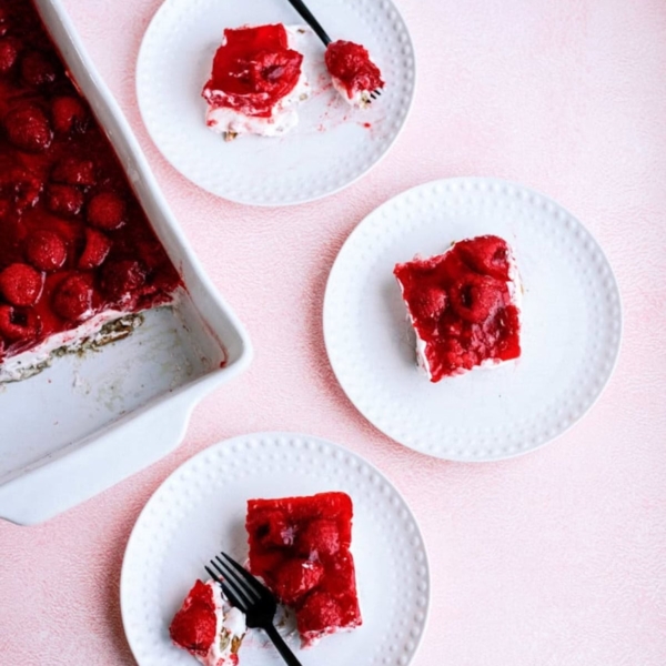 Three white plates with slices of raspberry dessert topped with red jelly, next to a baking dish containing the remaining dessert. Two plates have black forks.
