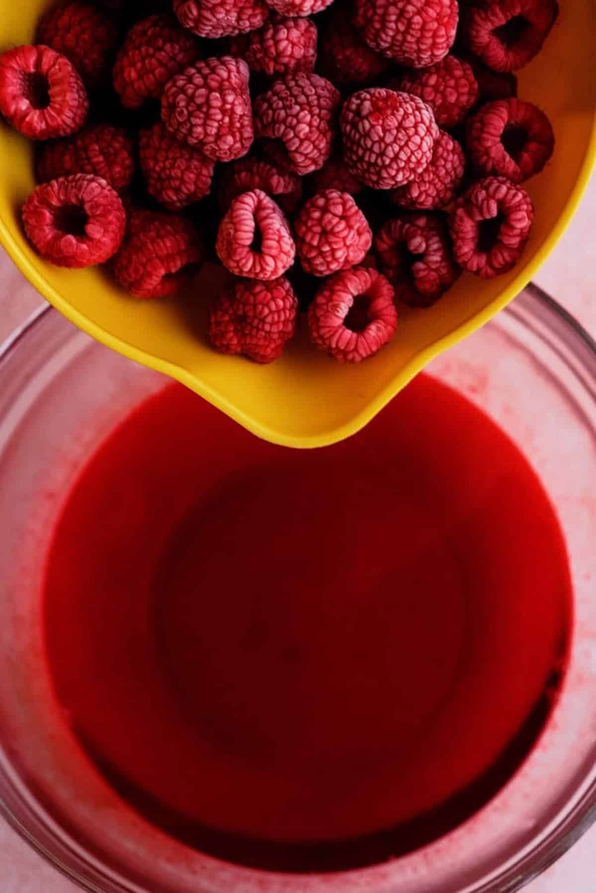 A yellow bowl of frozen raspberries is held above a glass bowl containing bright red raspberry juice.