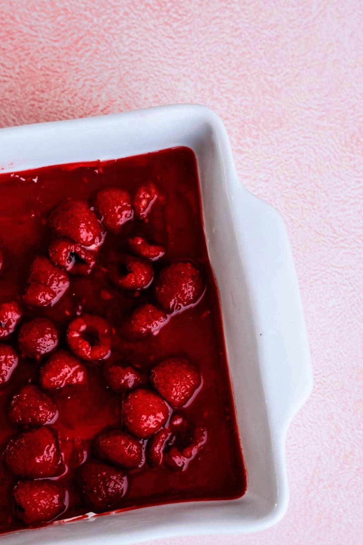 A white baking dish filled with whole raspberries in a thick red sauce sits on a pink textured surface.