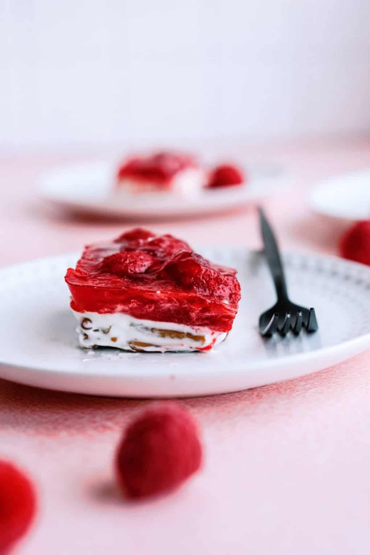 A slice of strawberry pretzel dessert with a fork on a white plate, topped with a red strawberry layer and creamy filling, with whole strawberries in the background.