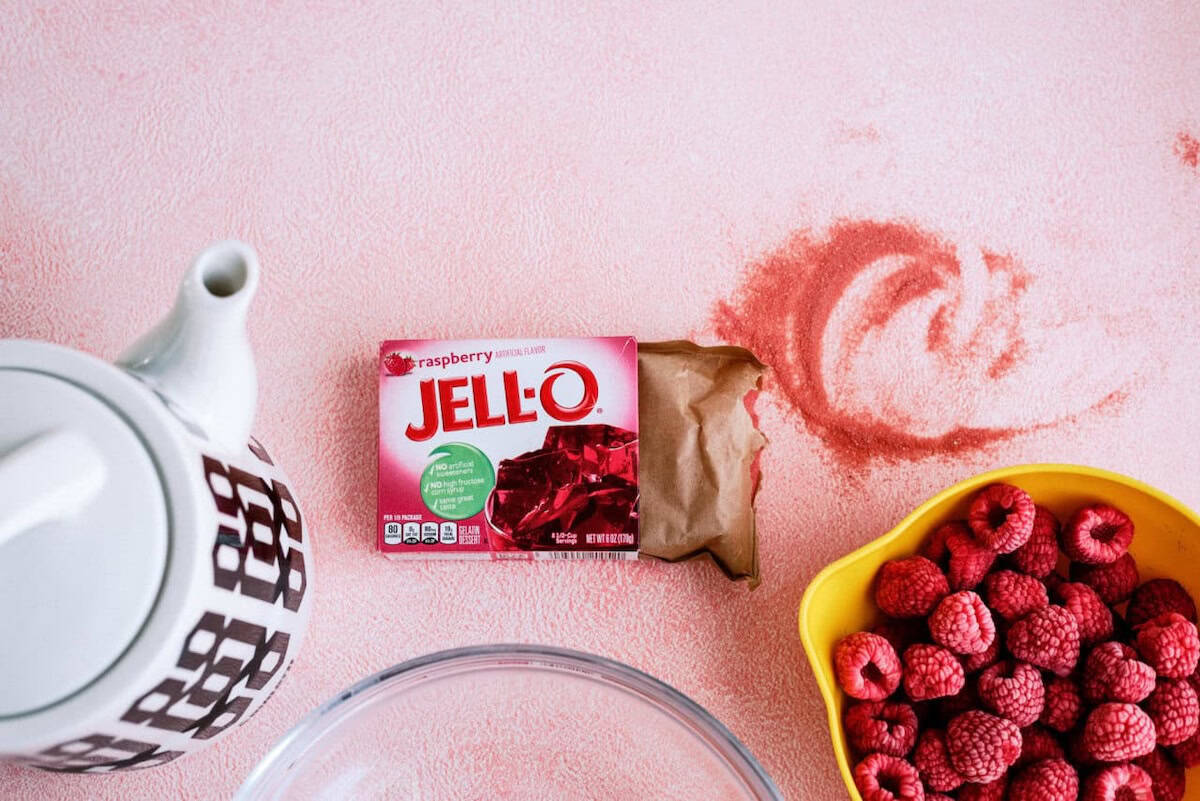 A box of raspberry Jell-O, a yellow bowl of raspberries, a teapot, and a glass bowl are arranged on a pink surface.