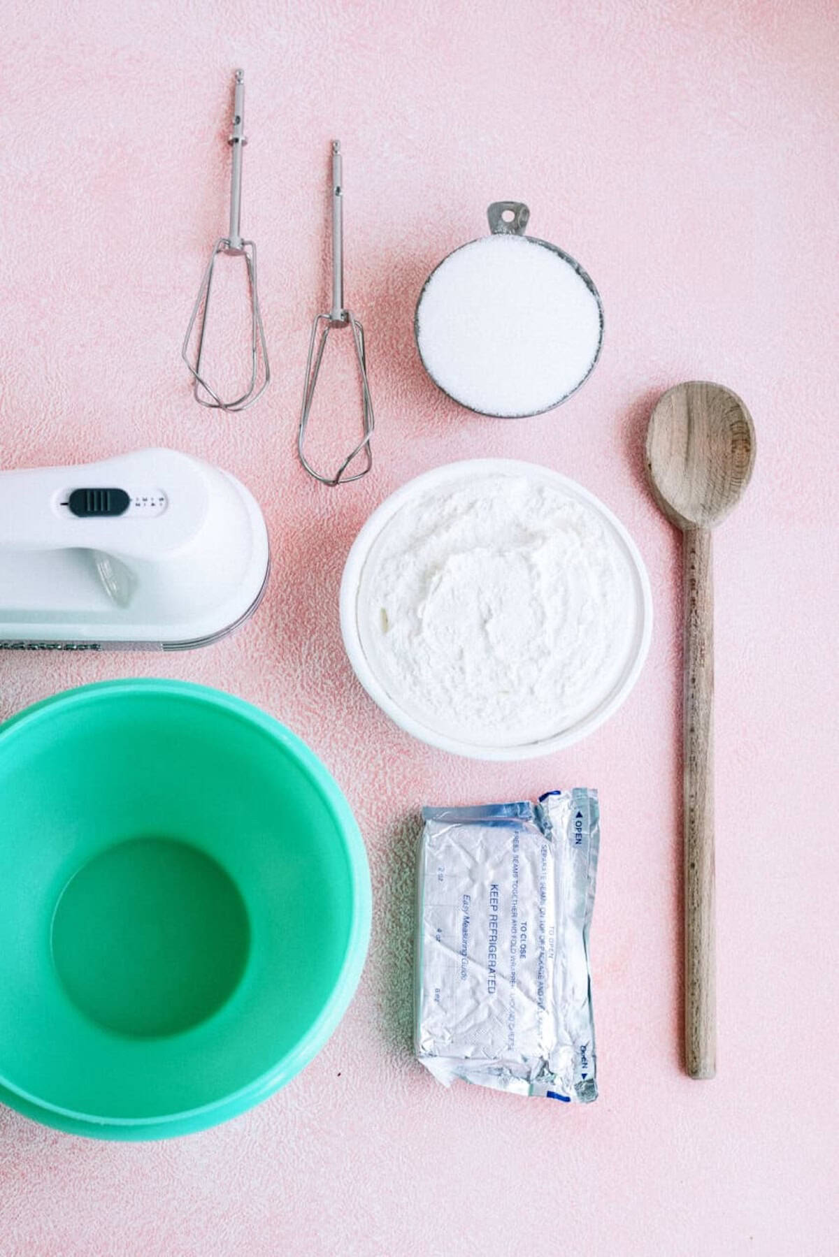 A person pours melted butter into a green bowl filled with cut green beans on a pink surface, with a white cloth, utensils, and scattered crumbs nearby.