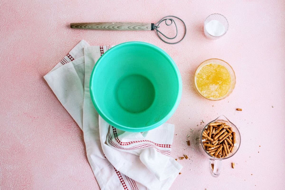 A green mixing bowl, a whisk, a glass of melted butter, a measuring cup with pellets, a small bowl of salt, and a white towel on a pink surface.