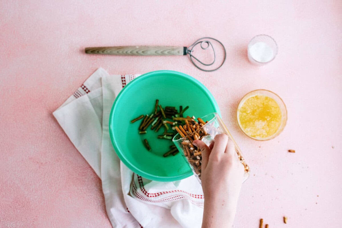 A hand pours pretzel sticks into a green bowl with green beans, next to melted butter, salt, a dough whisk, and a white kitchen towel on a pink surface.
