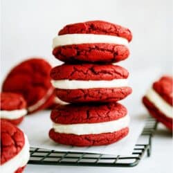 Red Velvet Whoopie Pie Sandwich Cookies stacked on a cooling rack lined with parchment paper with other Red Velvet Whoopie Pie Sandwich Cookies surrounding it.