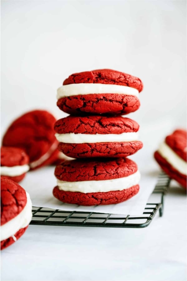 Red Velvet Whoopie Pie Sandwich Cookies stacked on a cooling rack lined with parchment paper with other Red Velvet Whoopie Pie Sandwich Cookies surrounding it.