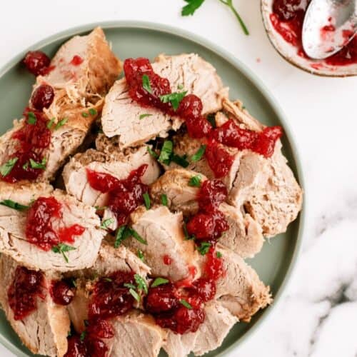 Sliced roast pork topped with cranberry sauce and chopped parsley on a green plate, with a small bowl of extra cranberry sauce and parsley on the side.