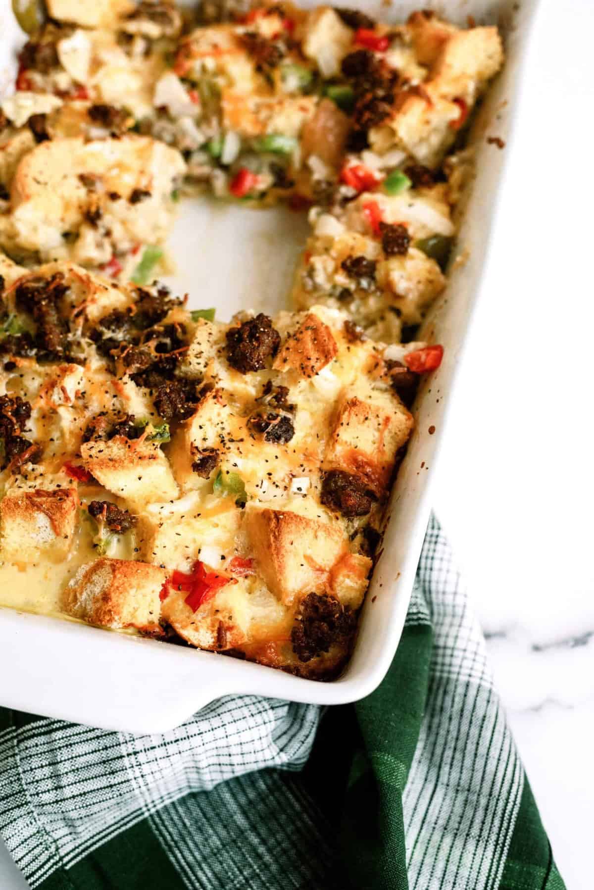 Baked casserole with bread cubes, bell peppers, and ground meat in a white dish on a green plaid cloth.