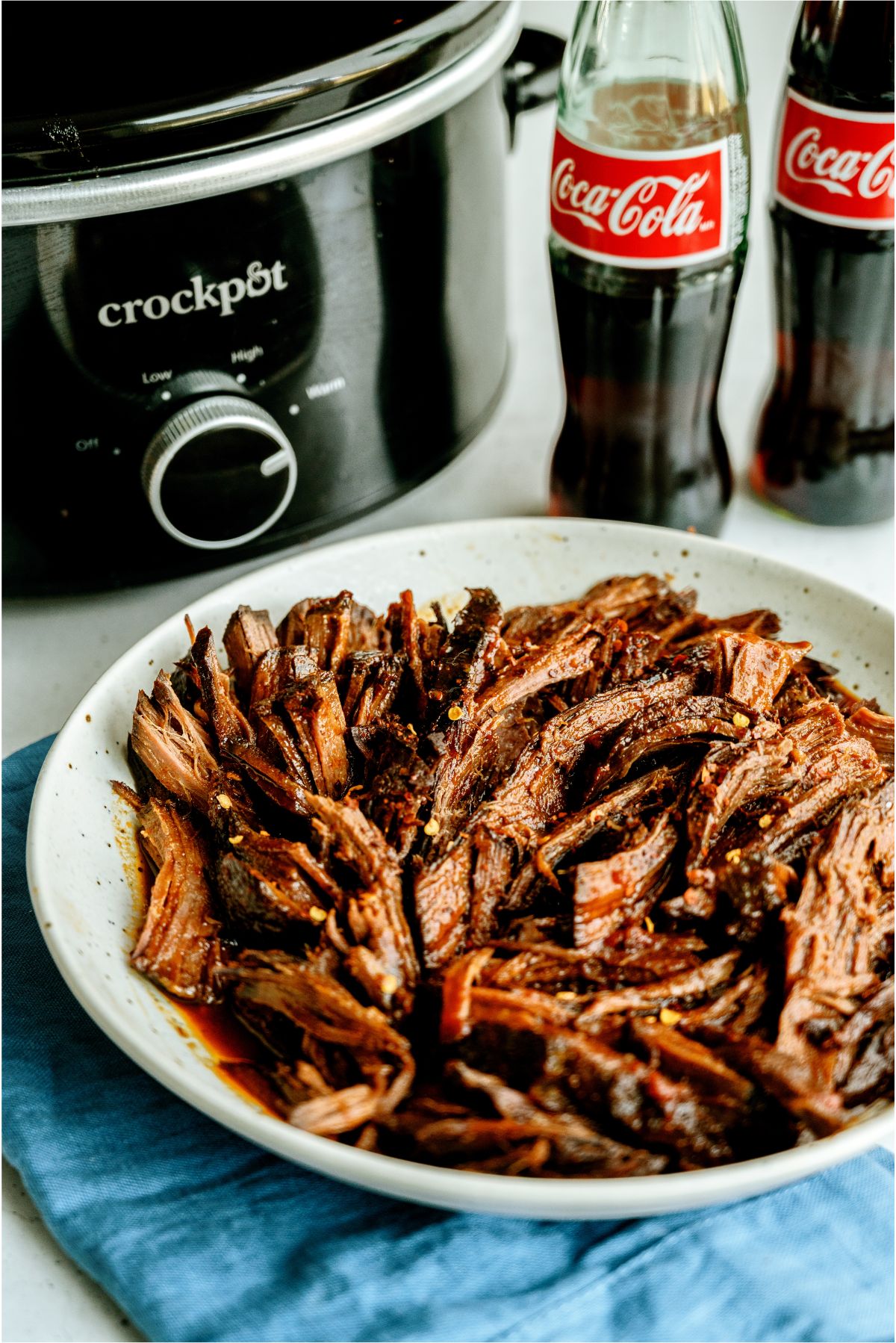 Slow Cooker Cola BBQ Roast Beef on a serving platter with a slow cooker and 2 bottles of coke in the background