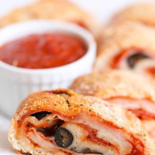 Close-up of a sliced stromboli filled with cheese, black olives, and pepperoni, served on a plate with a small bowl of marinara sauce in the background.