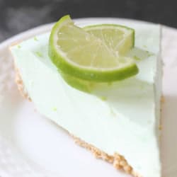 A slice of light green key lime pie with a graham cracker crust, garnished with two lime slices, served on a white plate.