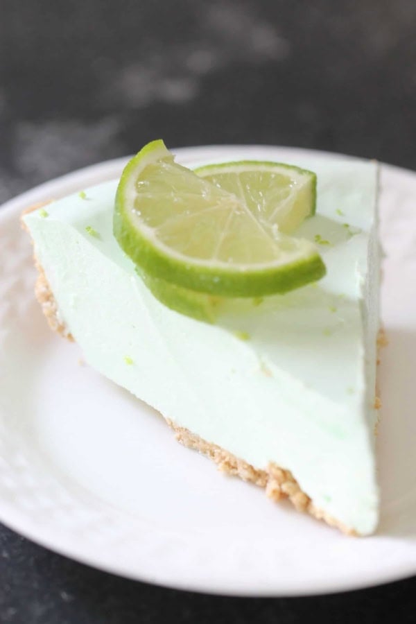 A slice of light green key lime pie with a graham cracker crust, garnished with two lime slices, served on a white plate.