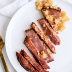 Sliced cooked ham topped with pineapple chunks arranged on a white plate, next to a gold fork and knife on a white surface.